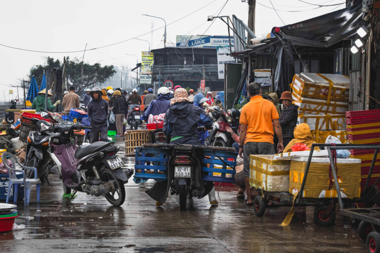 Mercado flotante vietnamita con motos, vendedores y cestas de productos bajo lluvia