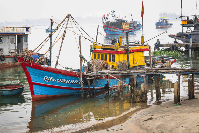 Barca pesquera roja y azul con velas amarillas en puerto vietnamita, otras embarcaciones y edificios al fondo