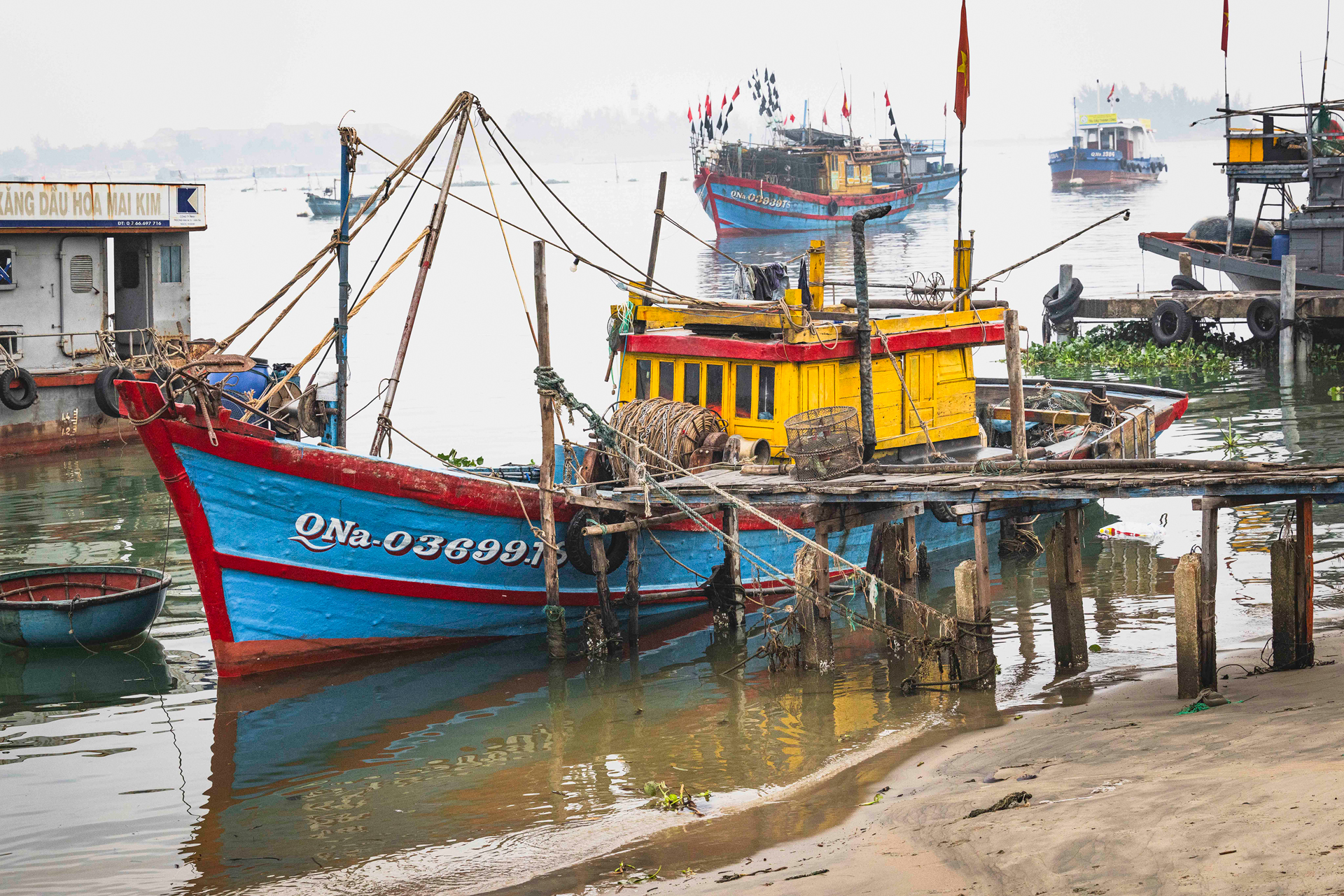 Barca pesquera roja y azul con velas amarillas en puerto vietnamita, otras embarcaciones y edificios al fondo