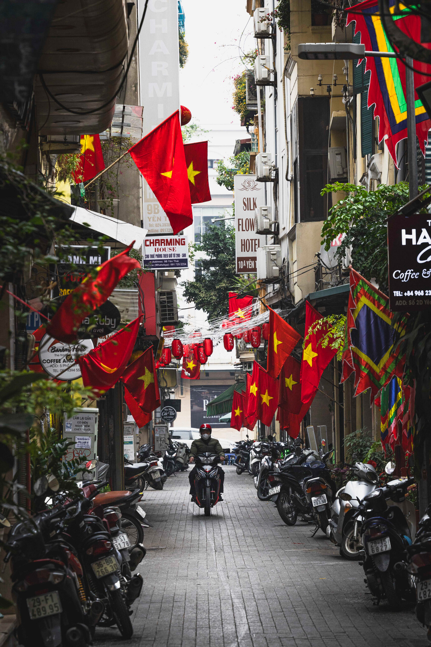 Callejuela vietnamita decorada con banderas rojas y amarillas, motos estacionadas a los lados, hoteles y cafeterías.