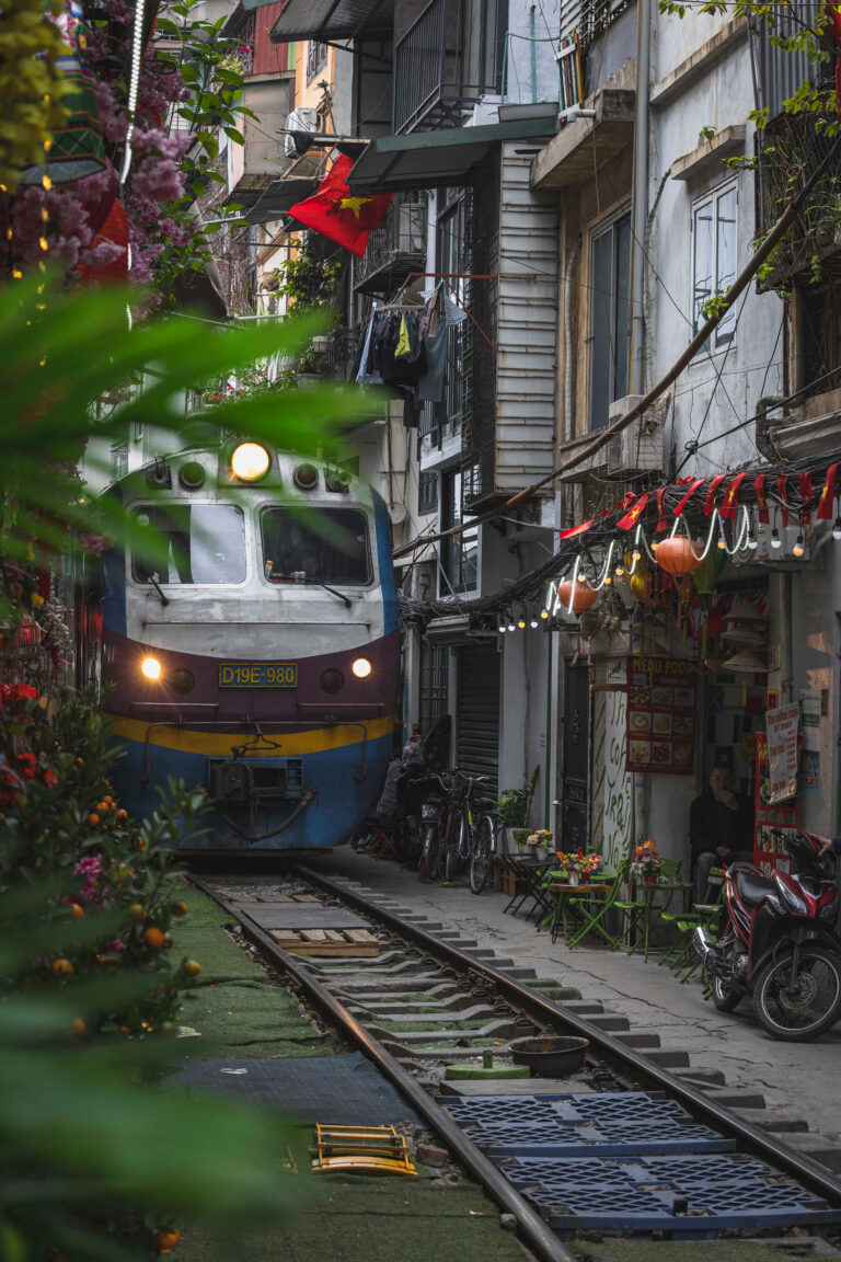 Tren atravesando un callejón estrecho entre edificios coloniales en Vietnam, rodeado de flores y vida urbana