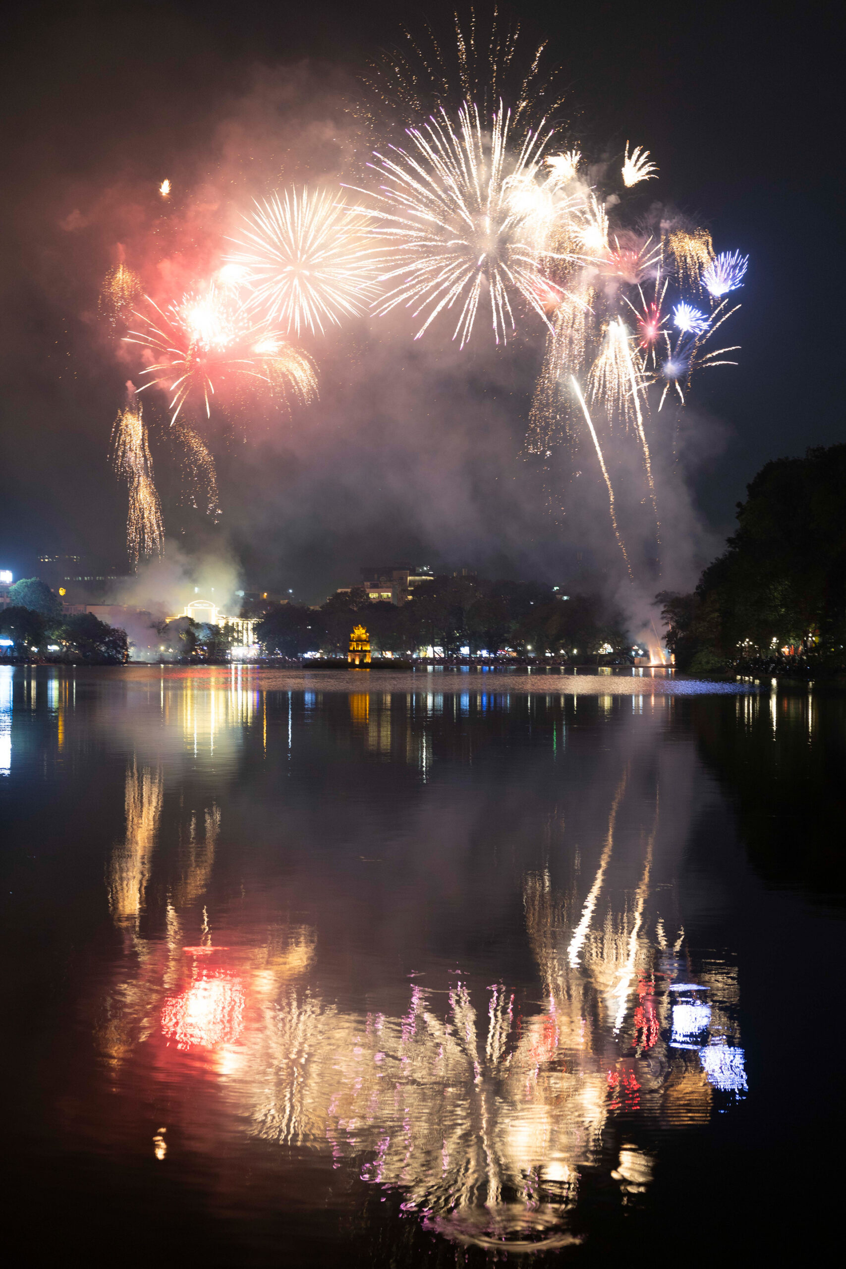 Fuegos artificiales reflejados en agua tranquila con edificios iluminados al fondo durante celebración nocturna