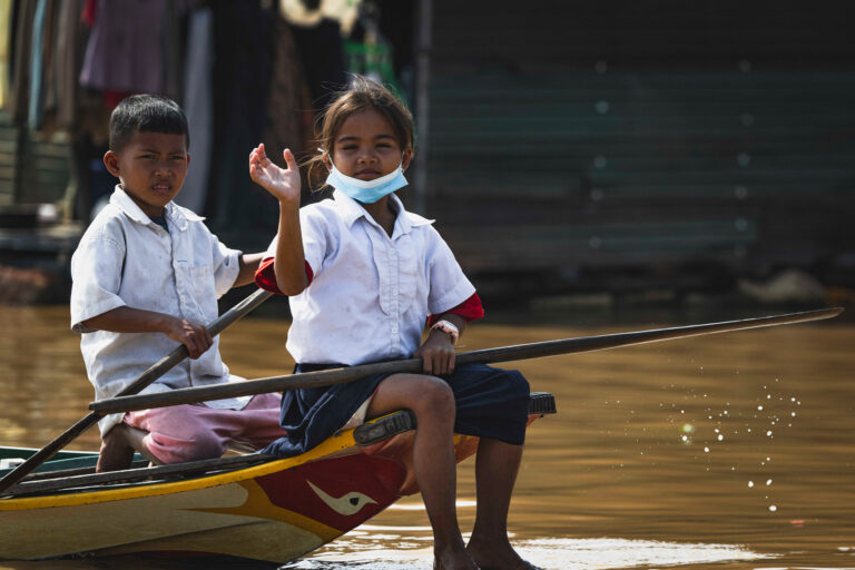 Dos niños en uniforme escolar blanco en una barca de madera en agua marrón, uno rema mientras sonríe a la cámara.