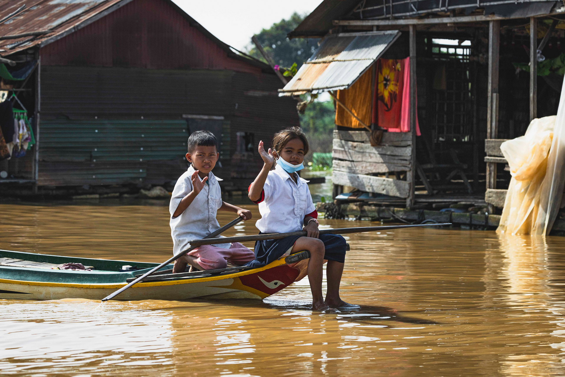 Dos niños en uniforme escolar en agua de inundación frente a casas de madera en Camboya