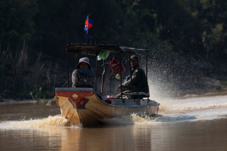 Bote tradicional con bandera camboyano navegando por río turbio con pasajeros a bordo