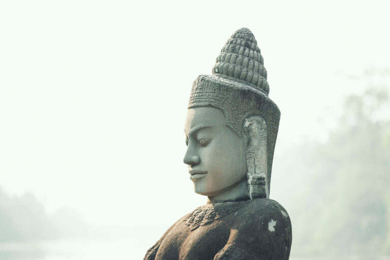 Estatua de piedra de Buda en perfil, con tocado puntiagudo y expresión serena contra cielo nublado