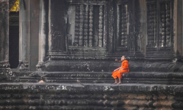 Monje budista en túnica naranja sentado en escalones de templo de piedra antigua en Camboya