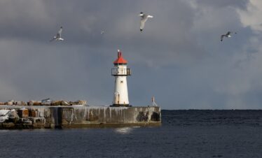 Faro blanco con torre roja en rompeolas de piedra, gaviotas volando sobre el mar gris