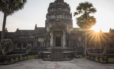 Templo de piedra con torre central al atardecer, rodeado de palmeras y estructuras antiguas en Camboya