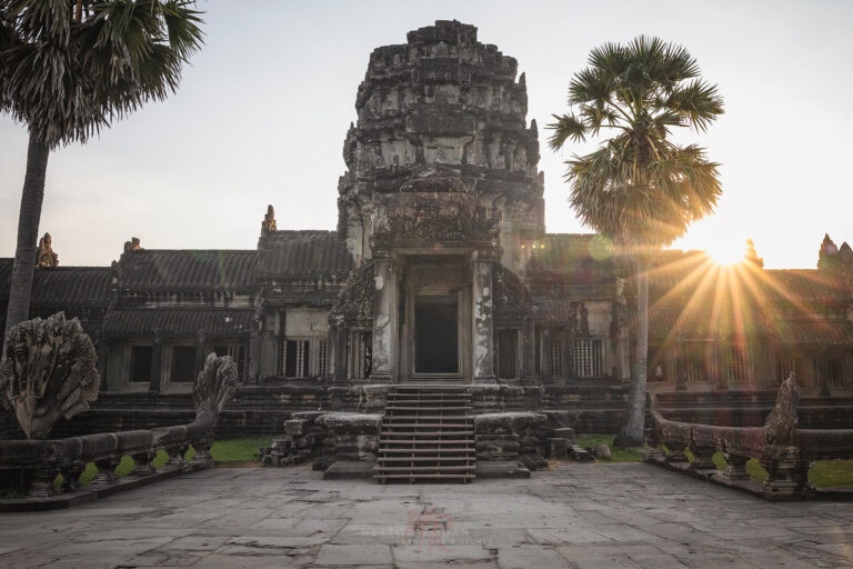 Templo de piedra con torre central al atardecer, rodeado de palmeras y estructuras antiguas en Camboya