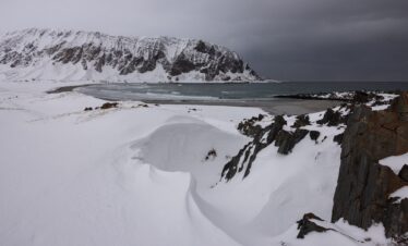 Playa nevada con montaña nevada al fondo y mar parcialmente congelado bajo cielo nublado