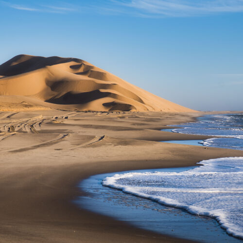 Duna de arena dorada junto a playa de olas azules en Namibia, con patrones de arena en primer plano
