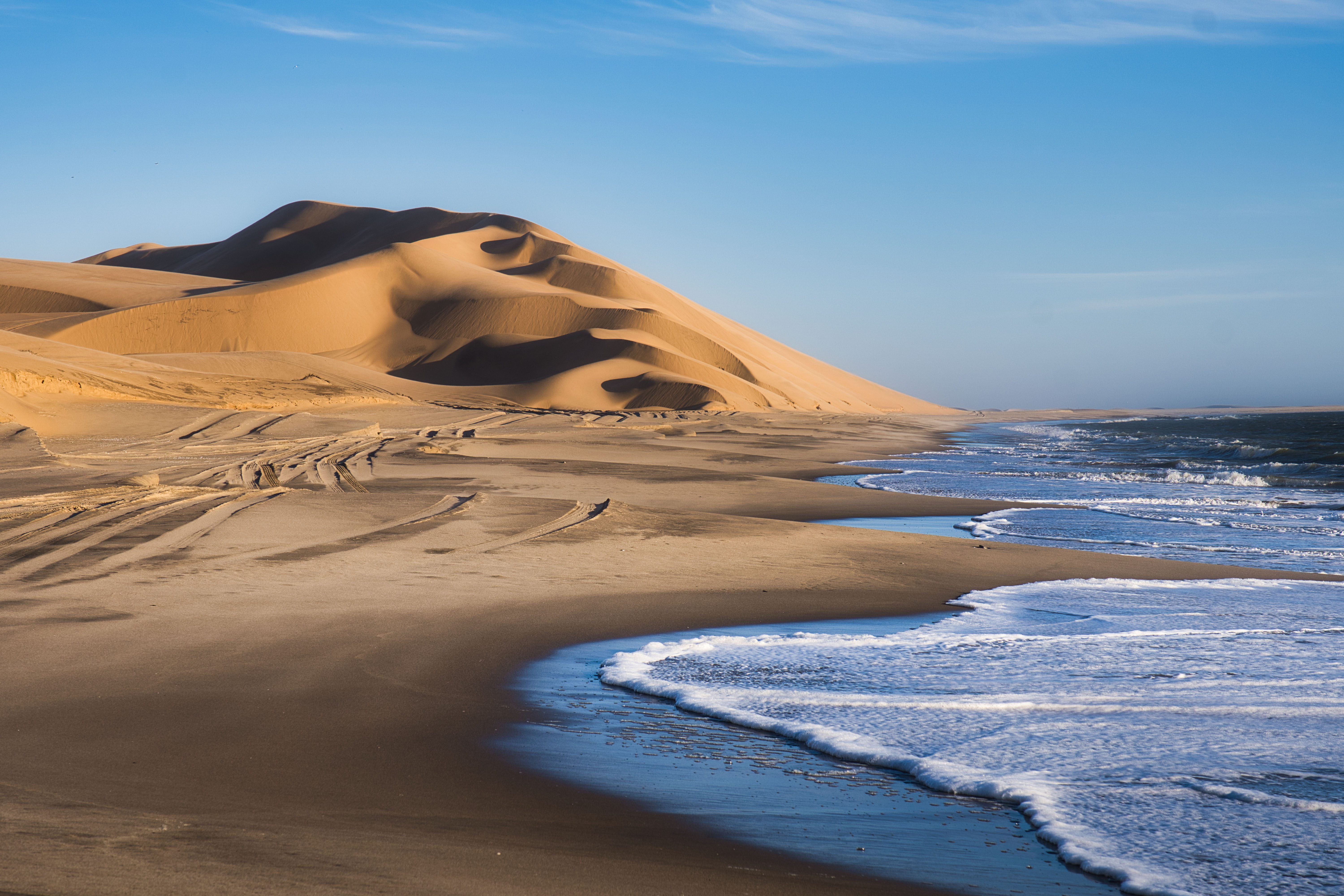 Duna de arena dorada junto a playa de olas azules en Namibia, con patrones de arena en primer plano