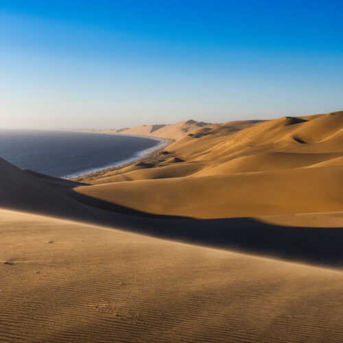 Dunas de arena dorada junto a océano azul en Namibia, con sombras marcadas y costa visible al fondo