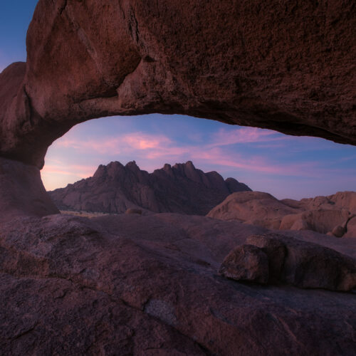 Arco natural de roca roja al atardecer en el desierto de Namibia, enmarcando montañas rocosas al fondo