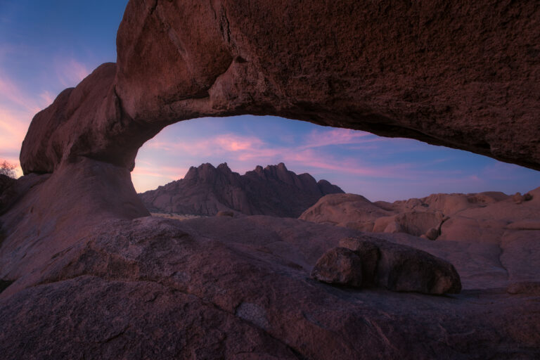 Arco natural de roca roja al atardecer en el desierto de Namibia, enmarcando montañas rocosas al fondo