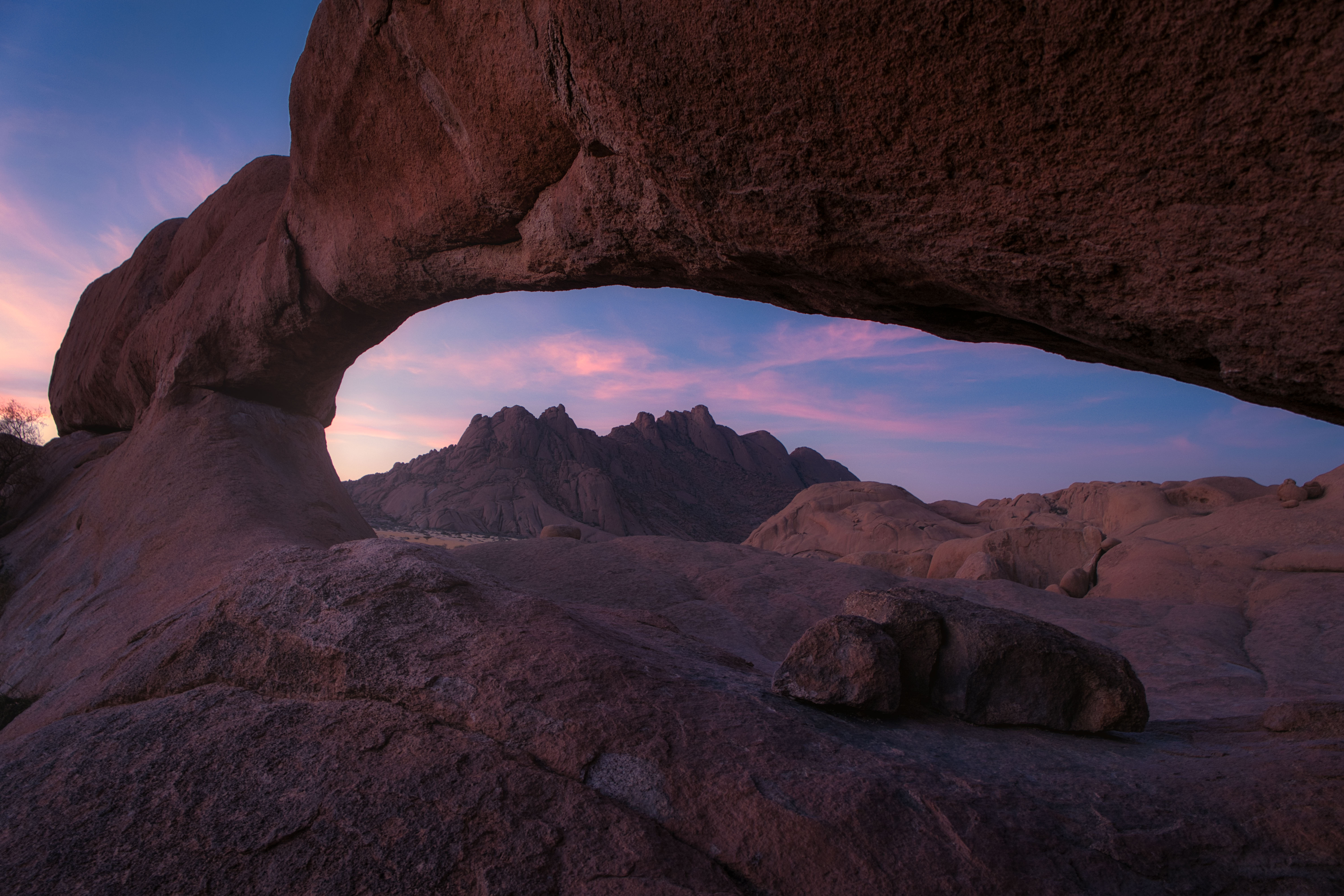 Arco natural de roca roja al atardecer en el desierto de Namibia, enmarcando montañas rocosas al fondo