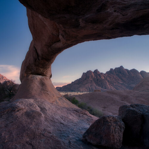 Formación rocosa con arco natural enmarcando montañas desérticas al atardecer en Namibia