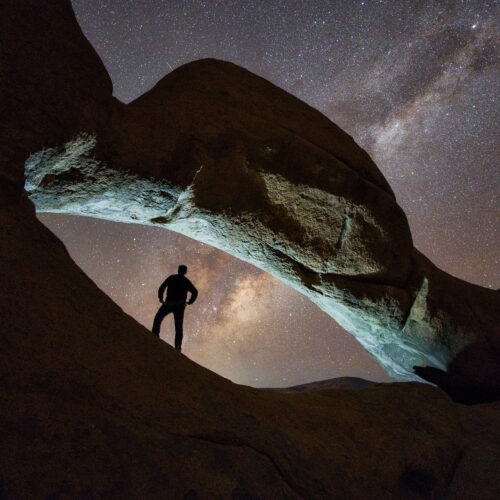 Viajero en silueta bajo formación rocosa natural con cielo estrellado y Vía Láctea visible en Namibia