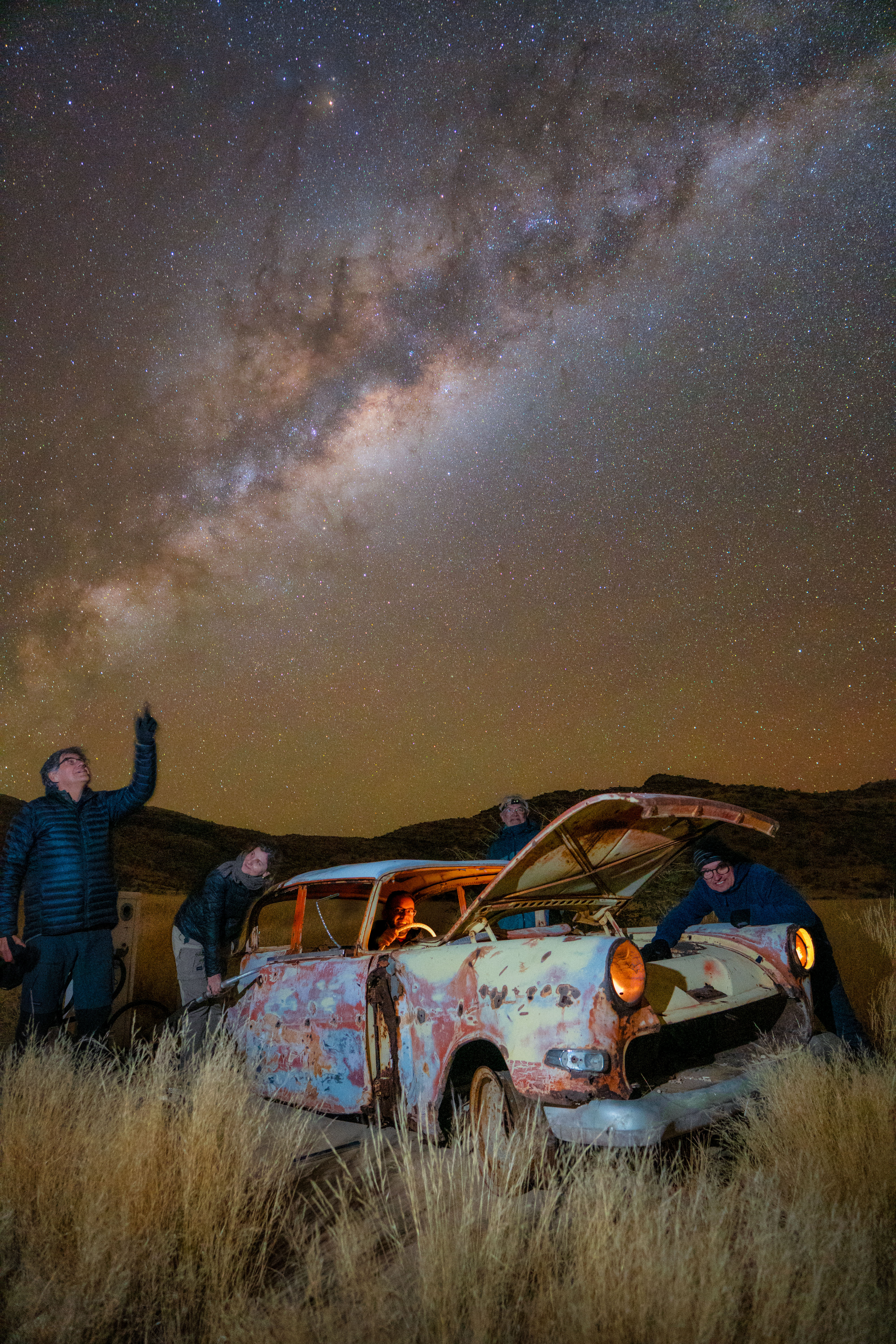 Grupo de viajeros junto a un vehículo abandonado bajo la Vía Láctea en el desierto de Namibia