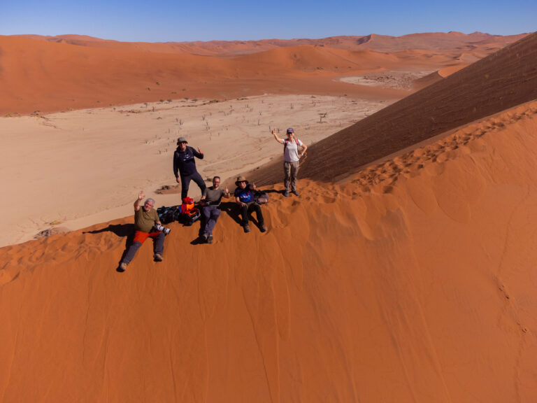 Grupo de aventureros en las dunas de arena roja de Namibia, saltando y posando en la ladera de una gran duna con valle desértico al fondo