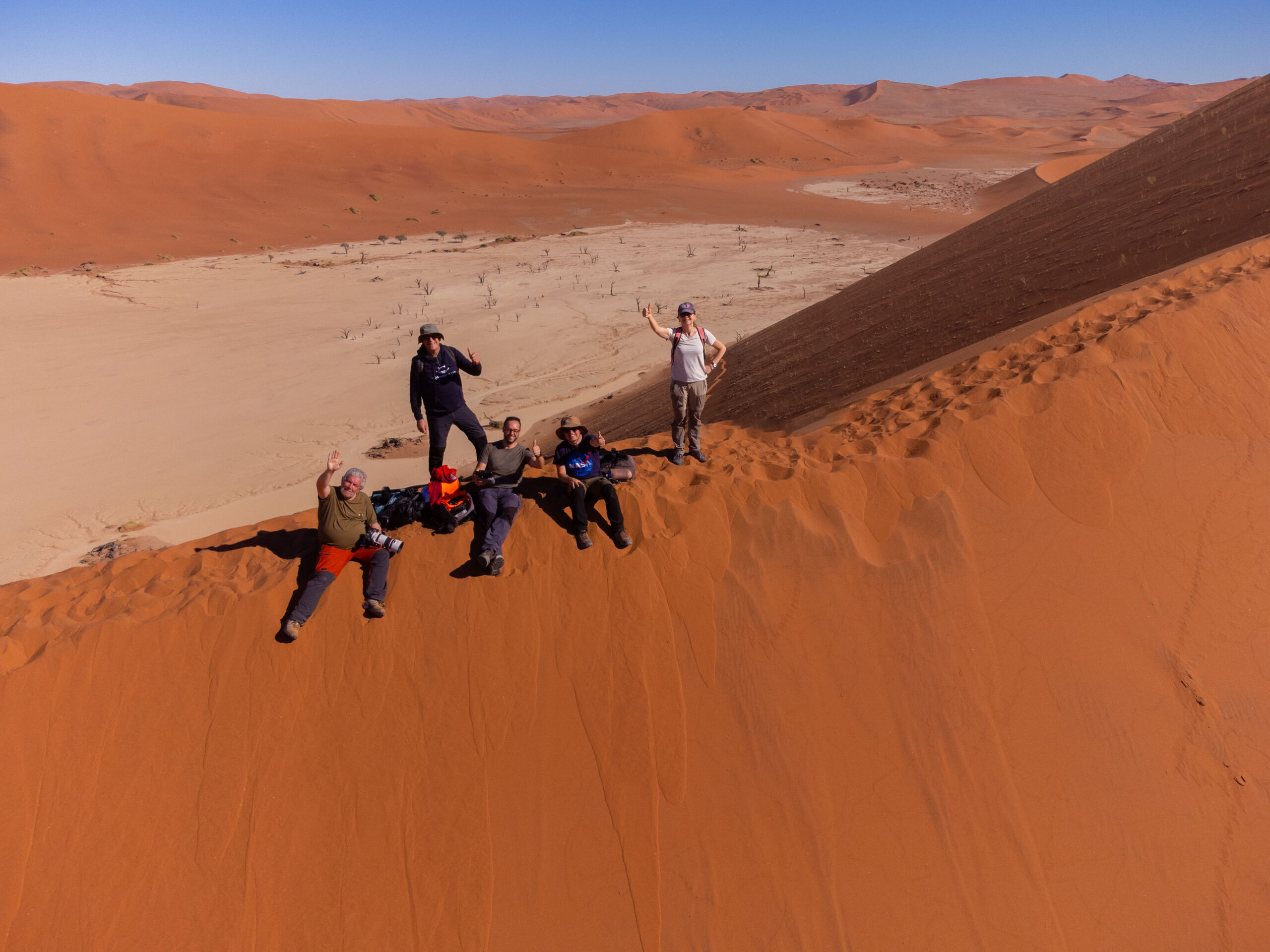 Grupo de aventureros en las dunas de arena roja de Namibia, saltando y posando en la ladera de una gran duna con valle desértico al fondo