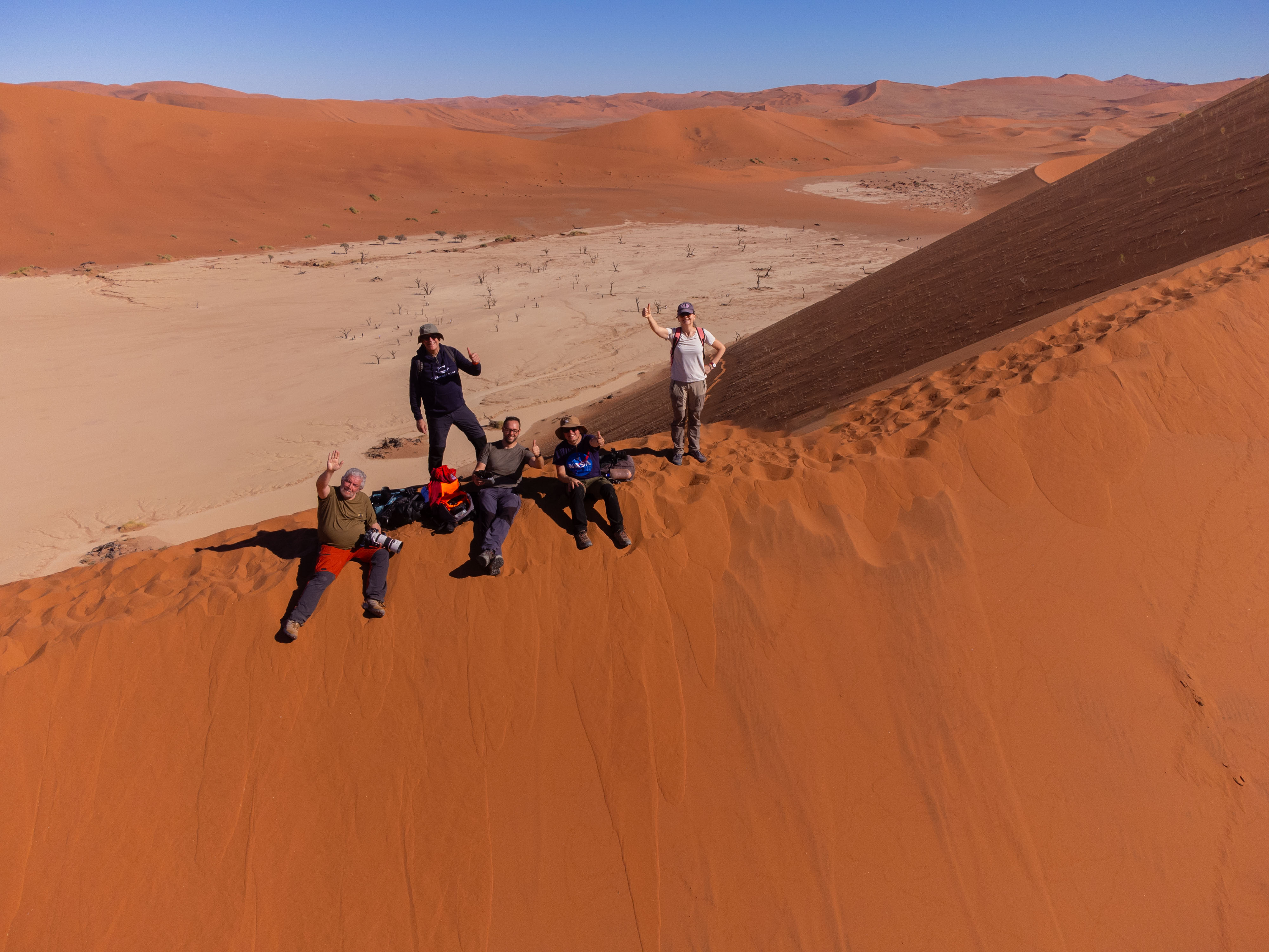Grupo de aventureros en las dunas de arena roja de Namibia, saltando y posando en la ladera de una gran duna con valle desértico al fondo