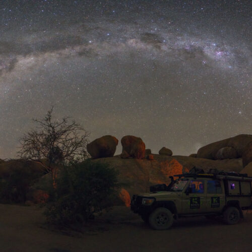 Campamento nocturno bajo la Vía Láctea en Spitzkoppe, Namibia, con vehículos todoterreno y fuego de campamento entre formaciones rocosas