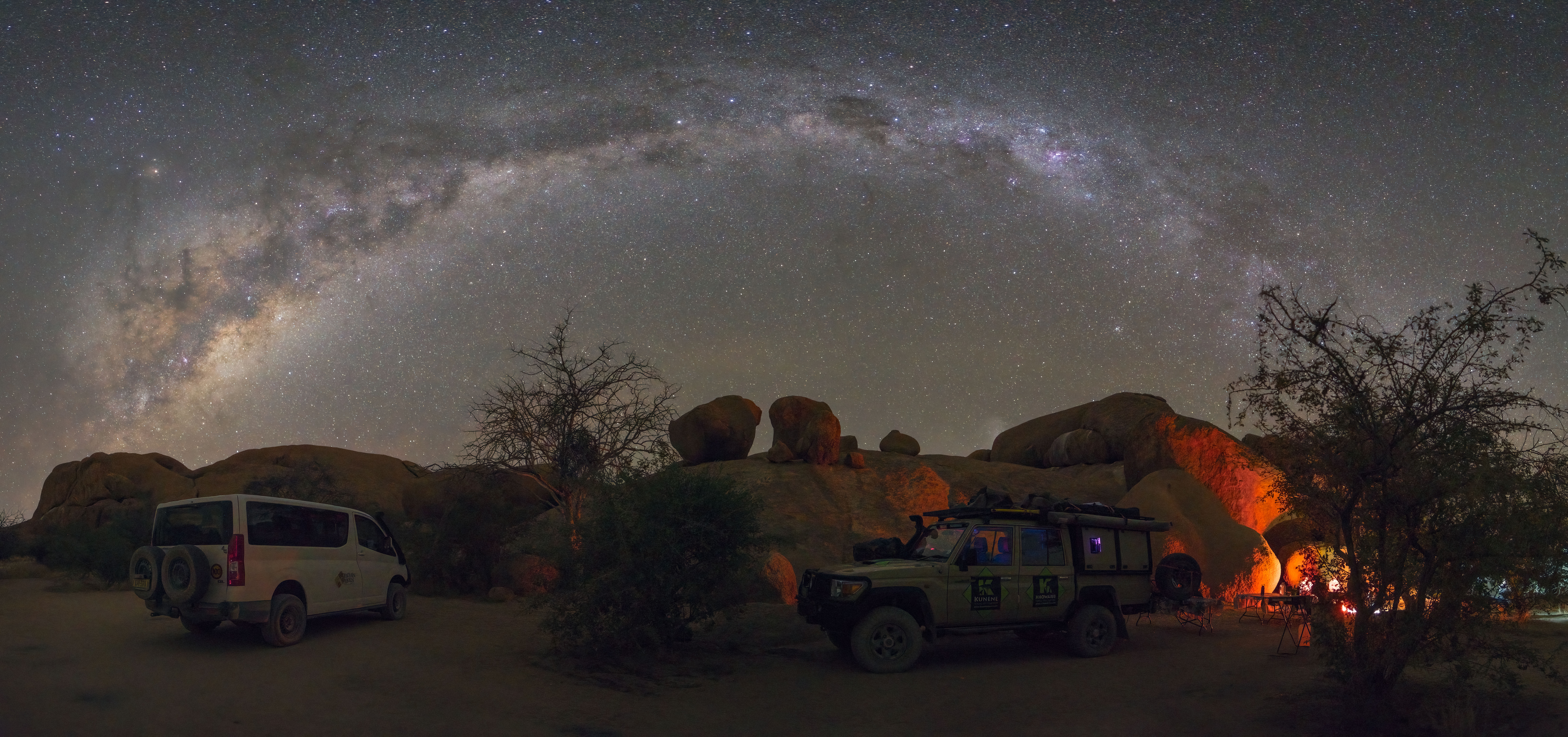 Campamento nocturno bajo la Vía Láctea en Spitzkoppe, Namibia, con vehículos todoterreno y fuego de campamento entre formaciones rocosas
