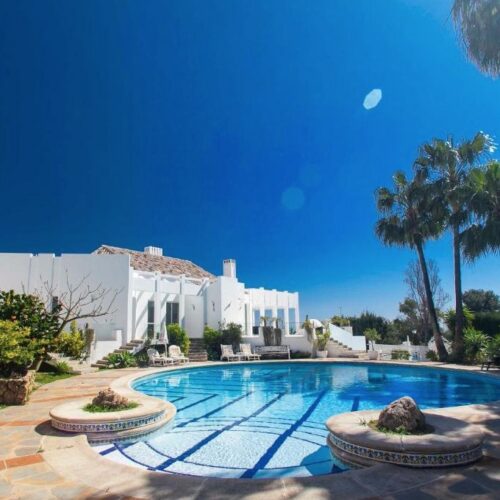 Villa blanca mediterránea con piscina olímpica azul, palmeras y cielo despejado