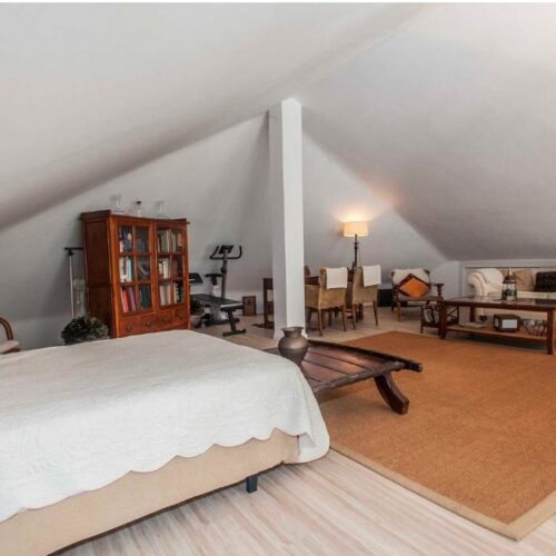 Loft amueblado en ático con cama, biblioteca, sofá y zona de estar bajo estructura de vigas blancas