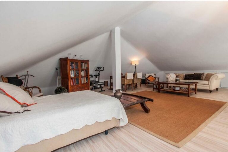 Loft amueblado en ático con cama, biblioteca, sofá y zona de estar bajo estructura de vigas blancas