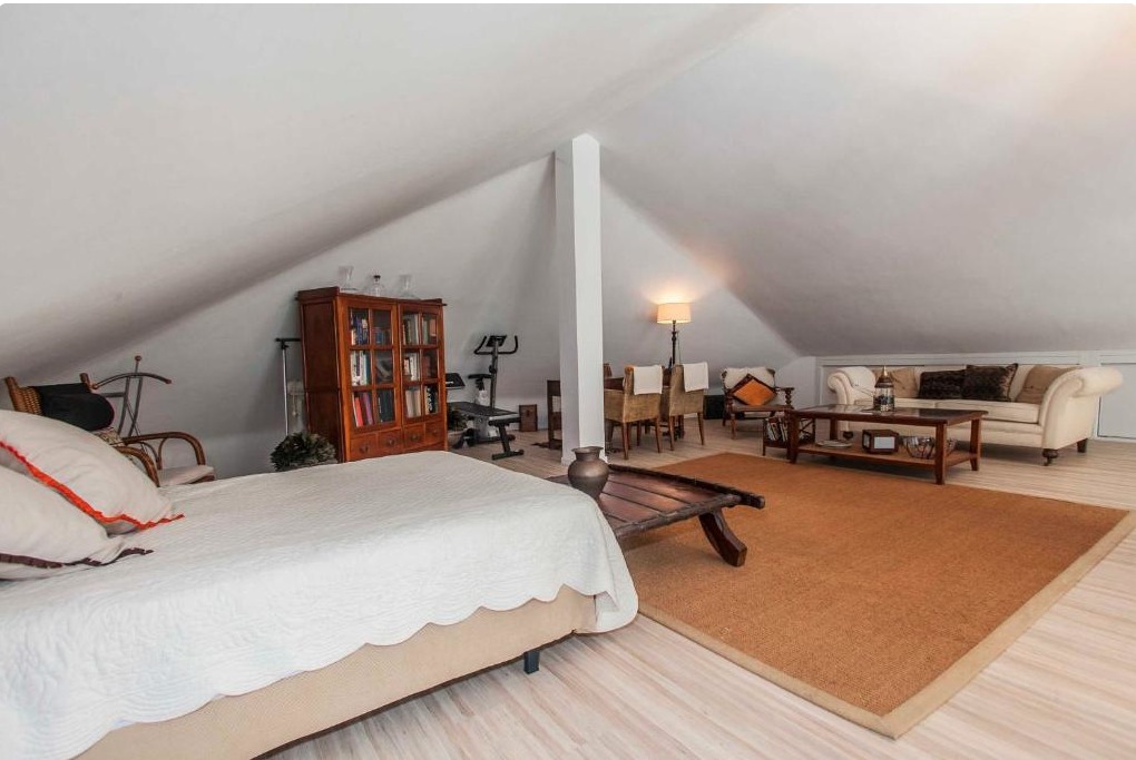 Loft amueblado en ático con cama, biblioteca, sofá y zona de estar bajo estructura de vigas blancas