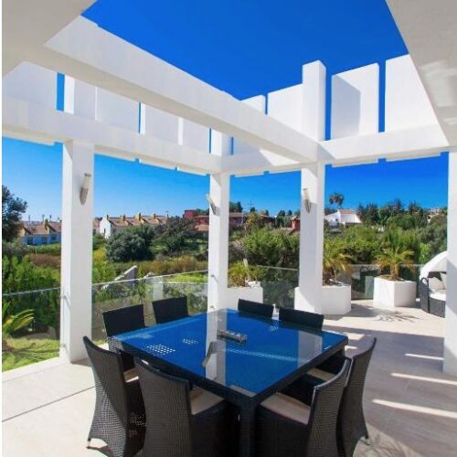Terraza de villa de lujo con toldo azul, mesa de comedor negra y vistas panorámicas a pueblo costero mediterráneo