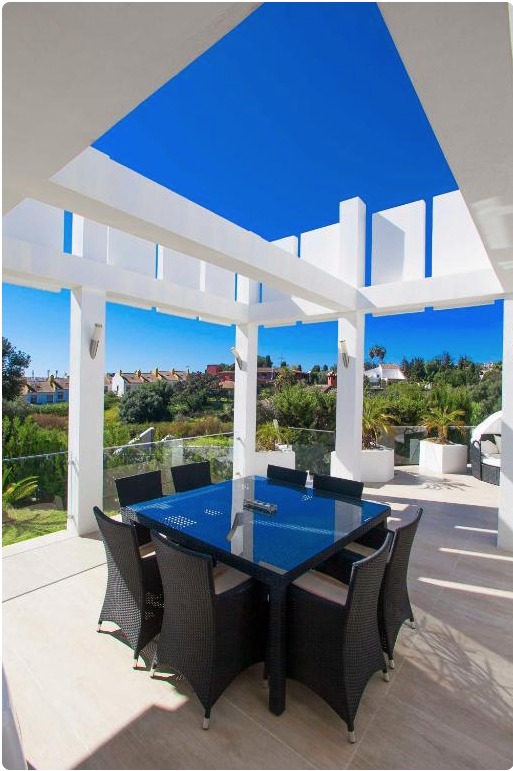 Terraza de villa de lujo con toldo azul, mesa de comedor negra y vistas panorámicas a pueblo costero mediterráneo