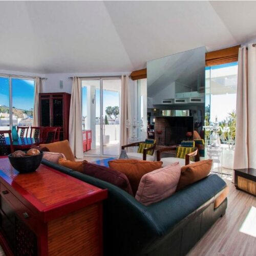 Luminoso salón de villa de lujo con sofá marrón, vista panorámica al mar Mediterráneo y terrazas acristaladas