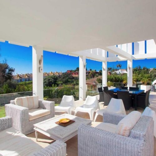 Terraza cubierta con pérgola blanca, muebles de ratán beige, vistas panorámicas a villa de lujo en Marbella y paisaje de montañas al atardecer