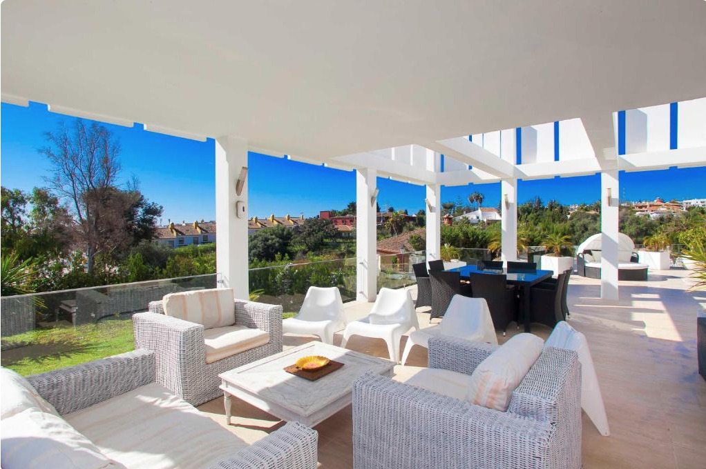 Terraza cubierta con pérgola blanca, muebles de ratán beige, vistas panorámicas a villa de lujo en Marbella y paisaje de montañas al atardecer