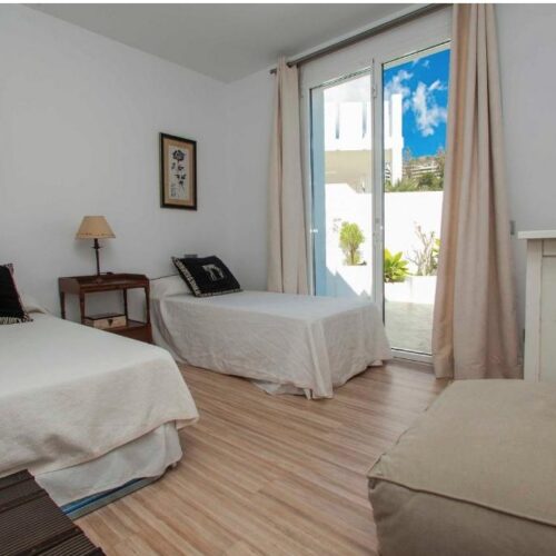 Dormitorio triple con tres camas individuales, suelo de madera, puertas correderas hacia terraza con vista al cielo azul y plantas