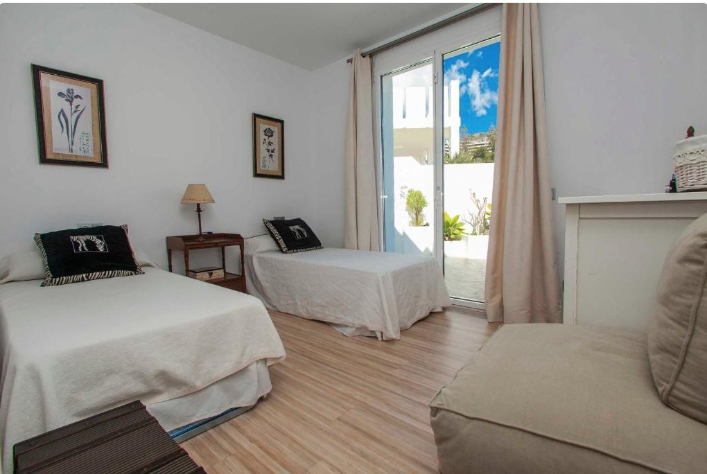 Dormitorio triple con tres camas individuales, suelo de madera, puertas correderas hacia terraza con vista al cielo azul y plantas