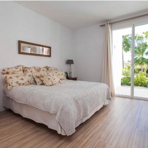 Dormitorio luminoso con cama doble, cabecero floral, mesita de noche de madera, lámpara y acceso a terraza con vistas al jardín tropical.