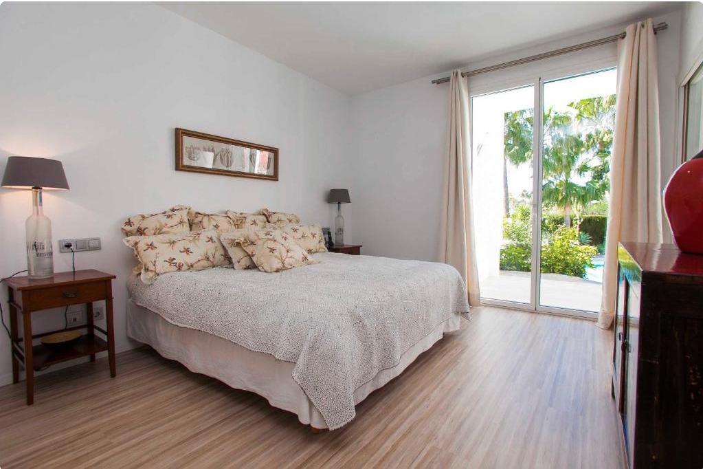 Dormitorio luminoso con cama doble, cabecero floral, mesita de noche de madera, lámpara y acceso a terraza con vistas al jardín tropical.