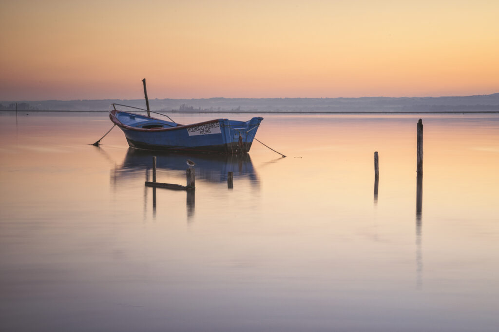 Barca de pesca azul anclada en aguas tranquilas al atardecer en la ría de Aveiro, con postes de amarre reflejados