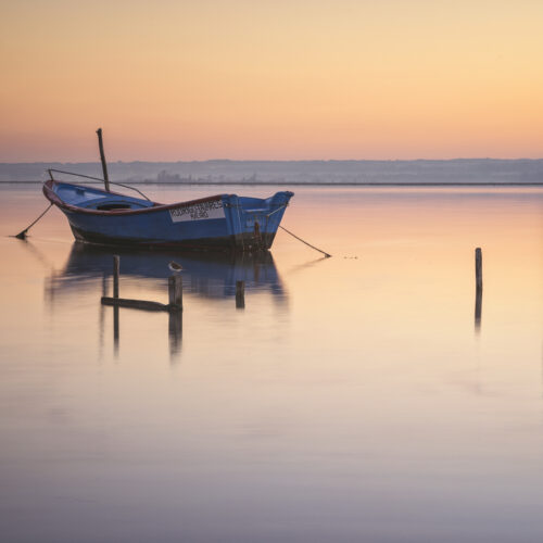 Barca de pesca azul anclada en aguas tranquilas al atardecer en la ría de Aveiro, con postes de amarre reflejados