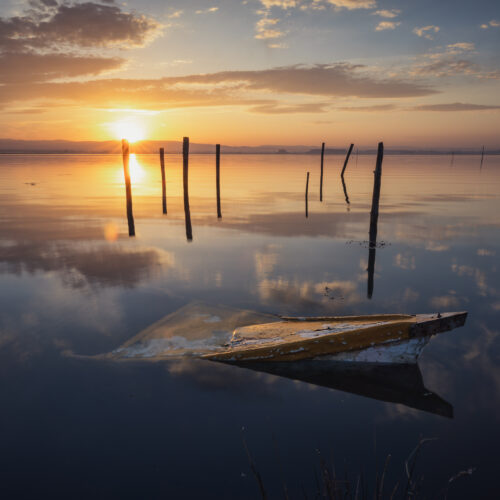 Barca de madera abandonada en aguas tranquilas al atardecer con postes de amarre reflejados