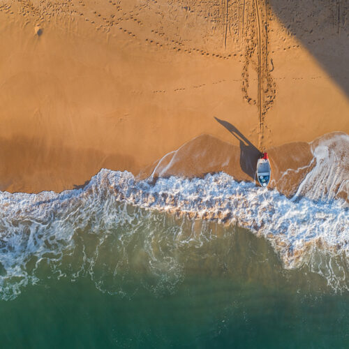Surfista con tabla en la orilla del Algarve, visto desde arriba con arena dorada y olas turquesas