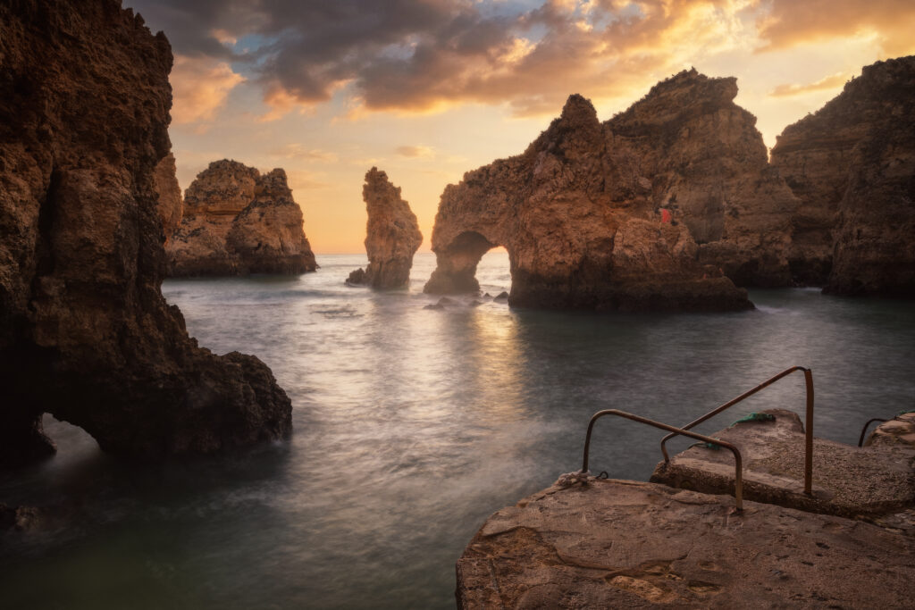 Acantilados rocosos del Algarve al atardecer con arco natural en el agua y escaleras de acceso en primer plano