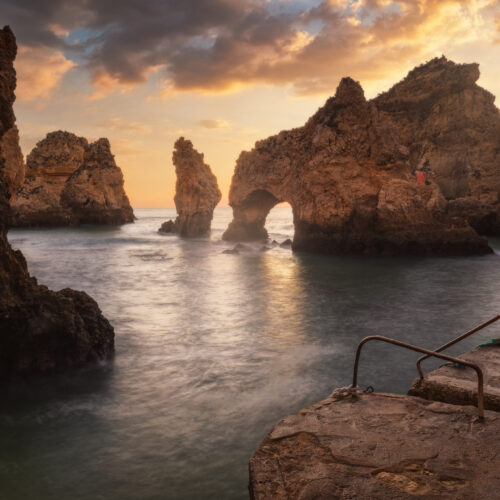 Acantilados rocosos del Algarve al atardecer con arco natural en el agua y escaleras de acceso en primer plano