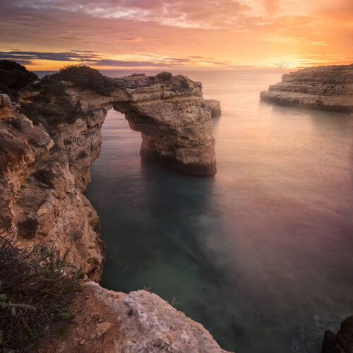 Arco de roca calcárea en acantilado del Algarve al atardecer con cielo naranja y mar en calma