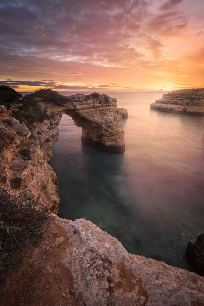 Arco de roca calcárea en acantilado del Algarve al atardecer con cielo naranja y mar en calma